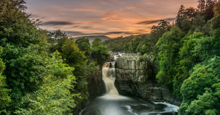 View of High Force Waterfall and surrounding countryside during sunset with pink sky.